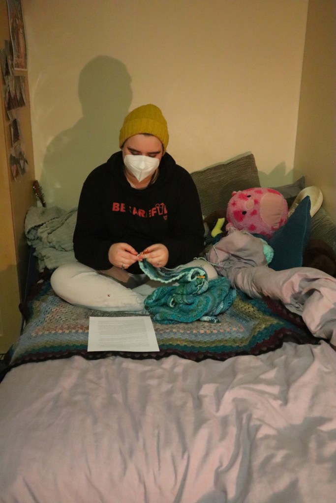 Aspen, a white masc person in a white face mask and blue beanie, sits crocheting on the bed.