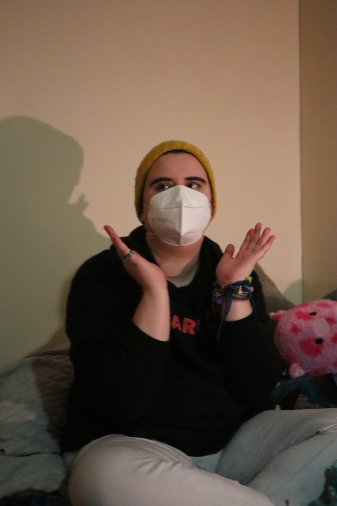 Aspen, wearing a black hoodie, white mask and yellow beanie, looking off camera and holding up their hands below their chin to illustrate the mask.