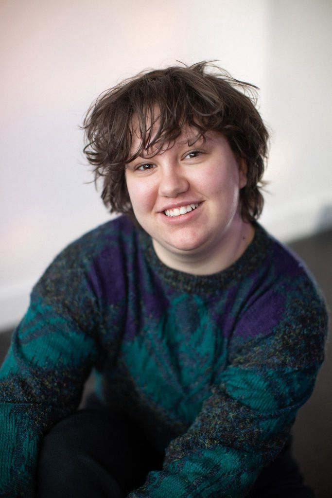 Quinn, a white androgynous person in a green jumper with brown hair, smiling at the camera.